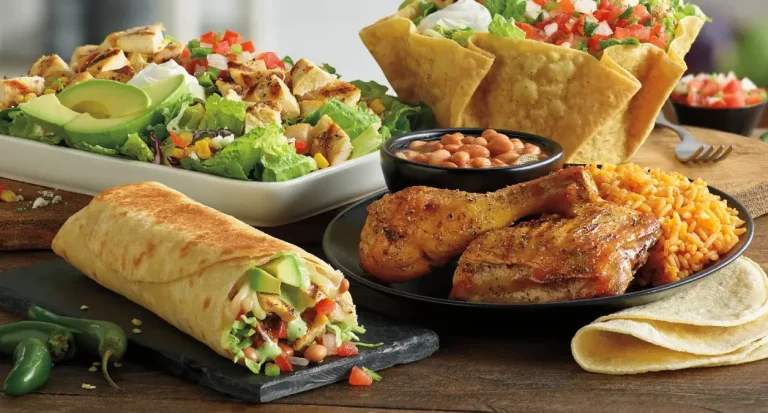 A spread of menu items on a wooden table, including a grilled chicken burrito, a chicken and rice plate with beans, tostada salad bowls, and a grilled chicken salad.