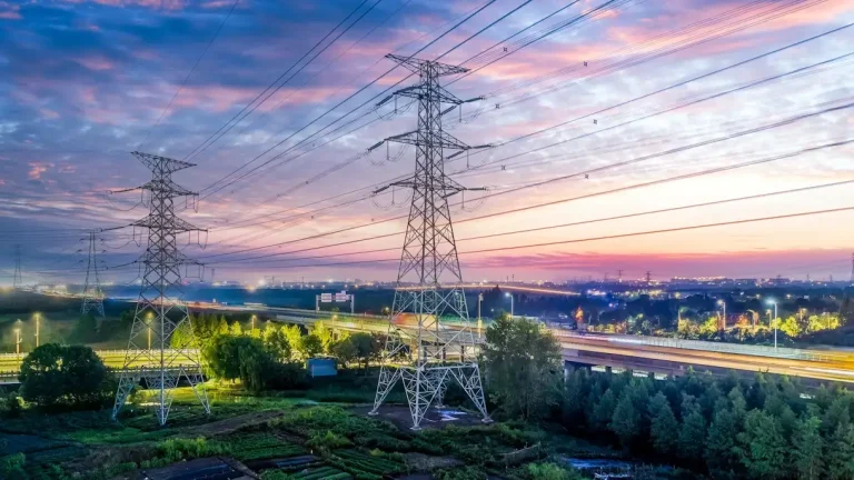 Aerial view of high-voltage electricity transmission towers and power lines at dusk, with a colorful sky and illuminated highway infrastructure in the background.